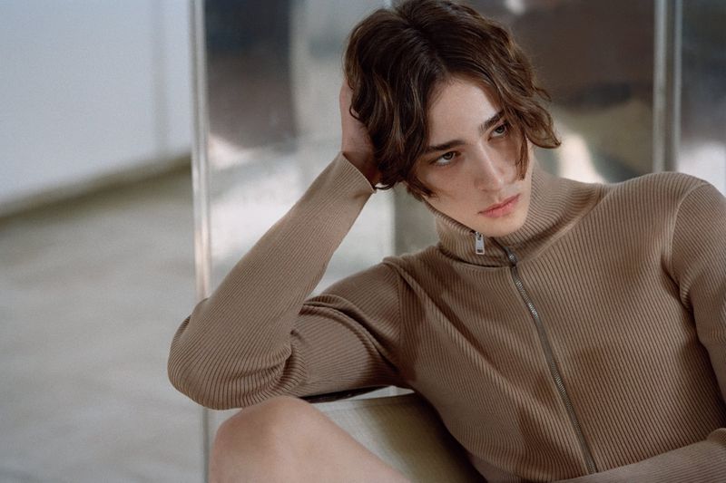 Portrait of a young adult with brown hair wearing a ribbed turtleneck posing indoors, looking off to the side with a neutral expression.