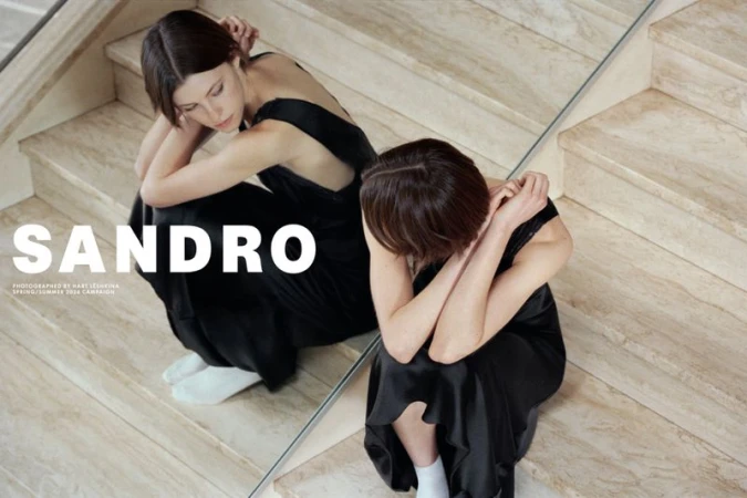 Two women wearing black dresses and white socks seated on marble stairs reflecting in a mirror for the Sandro campaign