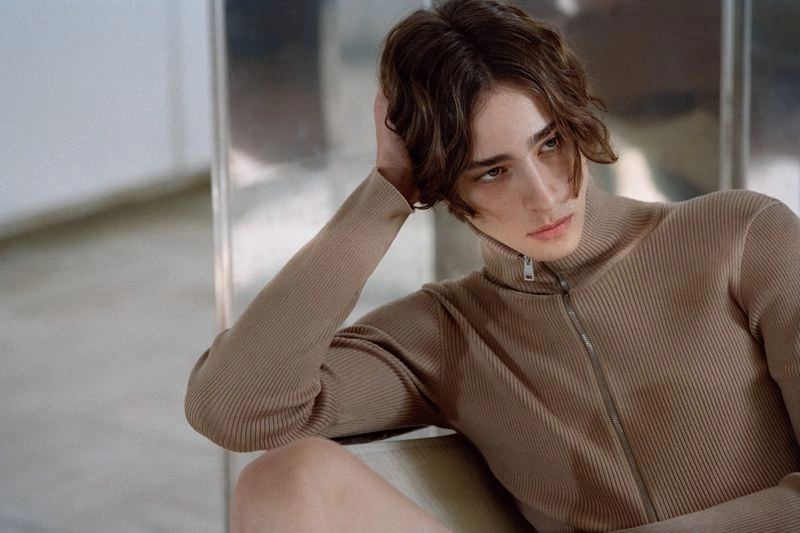 Close-up portrait of a male model wearing a ribbed camel-colored zip-up top with his hand behind his head.
