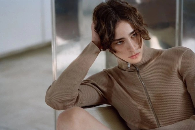 Close-up portrait of a male model wearing a ribbed camel-colored zip-up top with his hand behind his head.