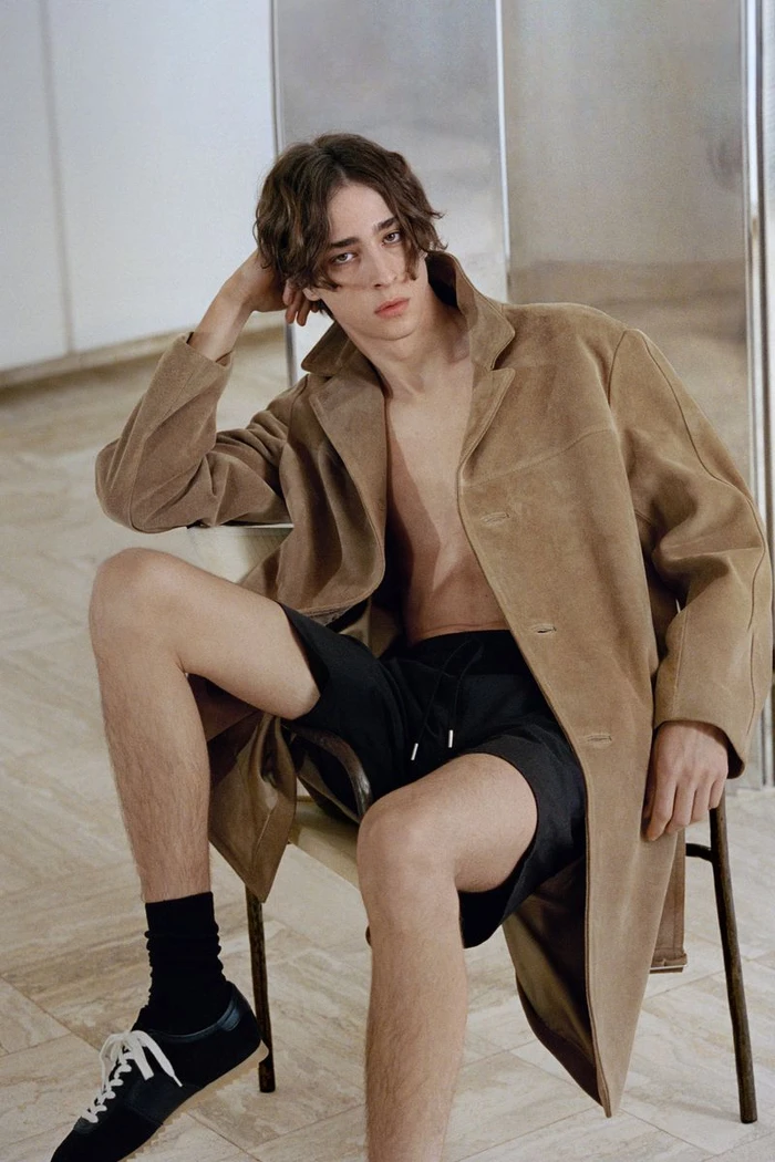Young male model seated on a chair wearing an open camel coat, black shorts, and sneakers against a minimalist indoor background.