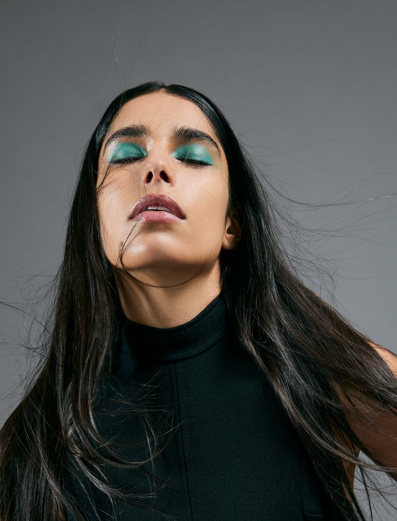Close-up of a brunette woman with her eyes closed, featuring dramatic green eyeshadow, glossy lips, and a black turtleneck top.