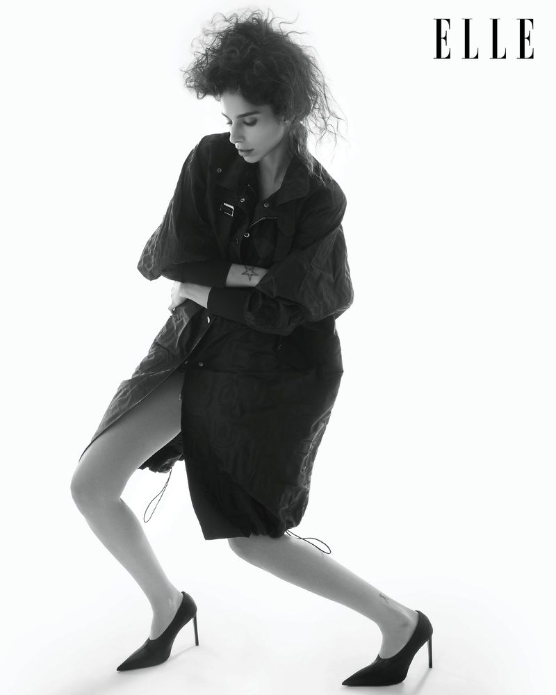 Fashion editorial shot of a slender adult woman posing in a black jacket and heels against a white background. Elle logo is visible.