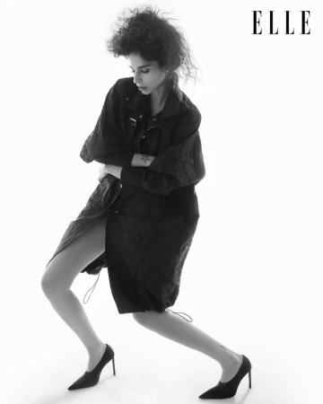 Fashion editorial shot of a slender adult woman posing in a black jacket and heels against a white background. Elle logo is visible.