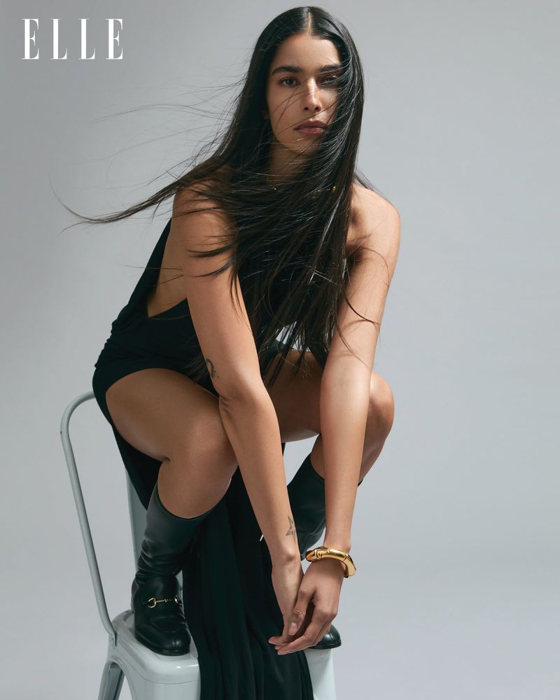 Fashion editorial shot of a woman with long dark hair, wearing a black dress and boots, seated on a white chair against a grey background.