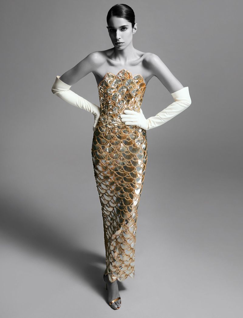 A fashion model poses in a gold sequin strapless gown with white gloves against a gray background in a studio fashion shot.