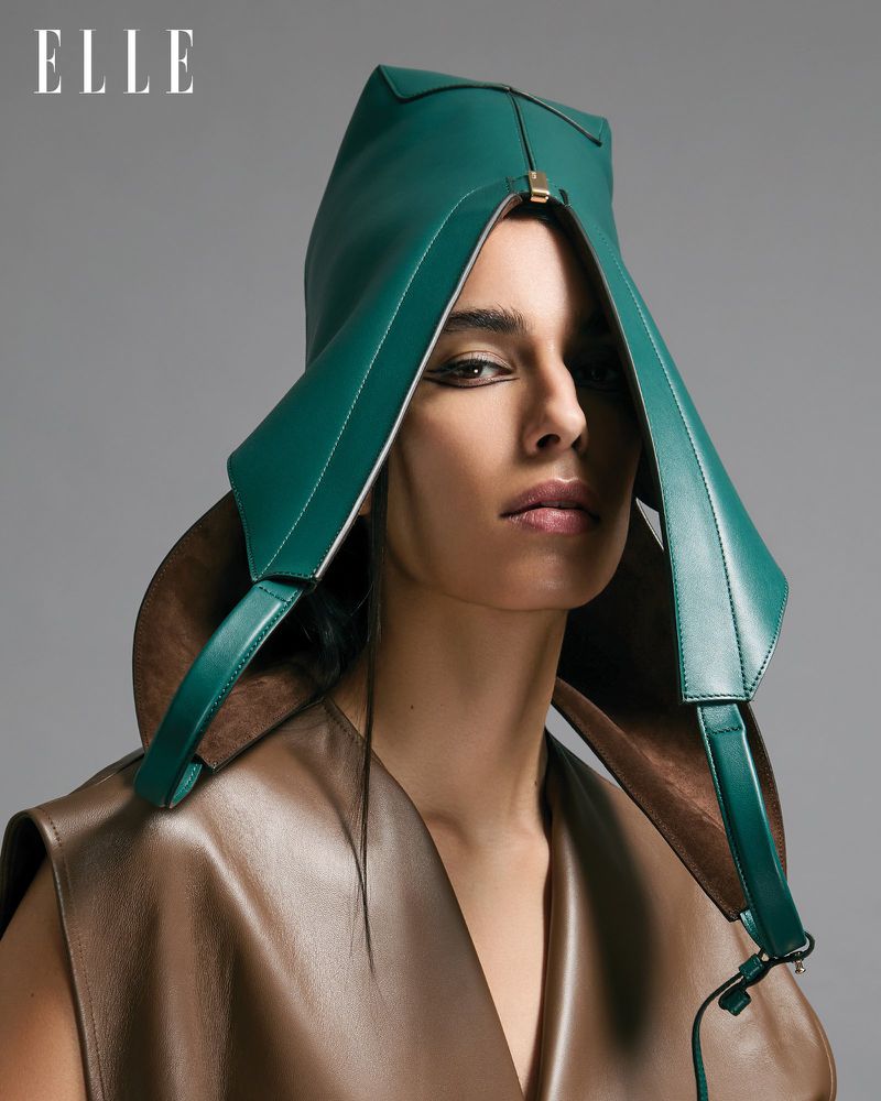 Fashion portrait of an adult woman wearing a green leather hood and brown leather top against a gray background in a photo studio.