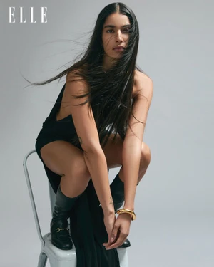 Model in a black dress and boots crouching on a white chair against a grey seamless background for Elle India