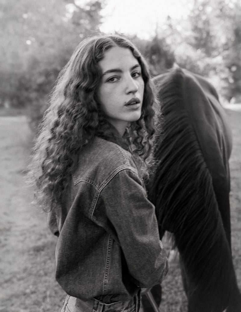Young woman with long curly hair wearing a denim jacket standing outdoors next to a dark horse in a black and white photograph.