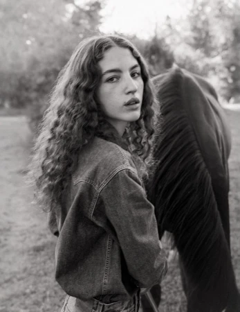 Young woman with long curly hair wearing a denim jacket standing outdoors next to a dark horse in a black and white photograph.