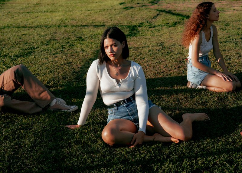 Three models relaxing on a sunny grass field wearing denim shorts and casual tops for Levi's campaign photography.