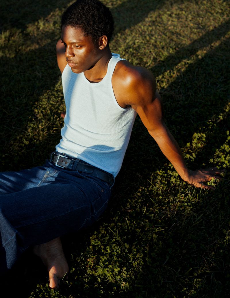Black man wearing a white tank top and blue denim jeans seated outdoors on a grassy lawn in sunlight.