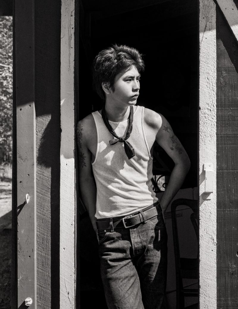 Young man in a white tank top and dark jeans leaning against a doorway in black and white