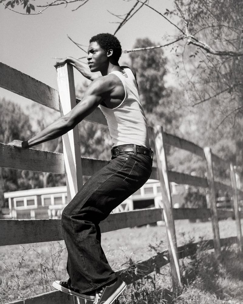 Black man wearing a white tank top and dark jeans climbing a wooden fence outdoors