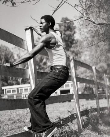 Black man wearing a white tank top and dark jeans climbing a wooden fence outdoors