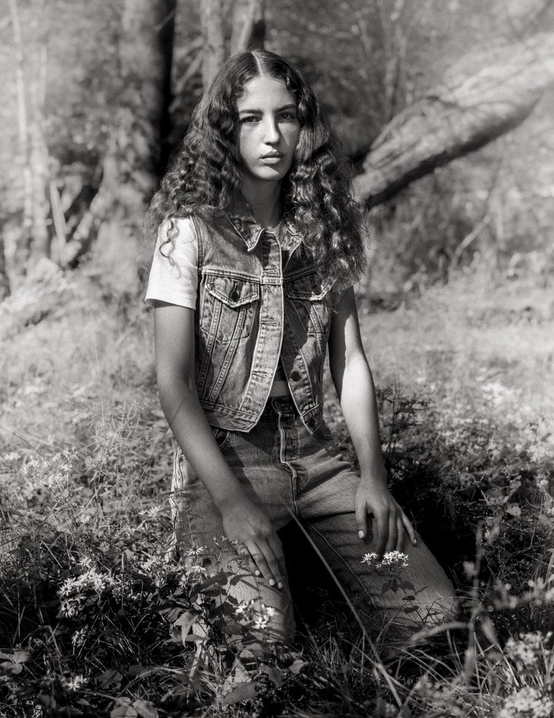 Model with long curly hair wearing a denim vest and jeans kneeling outdoors in a wooded meadow setting.