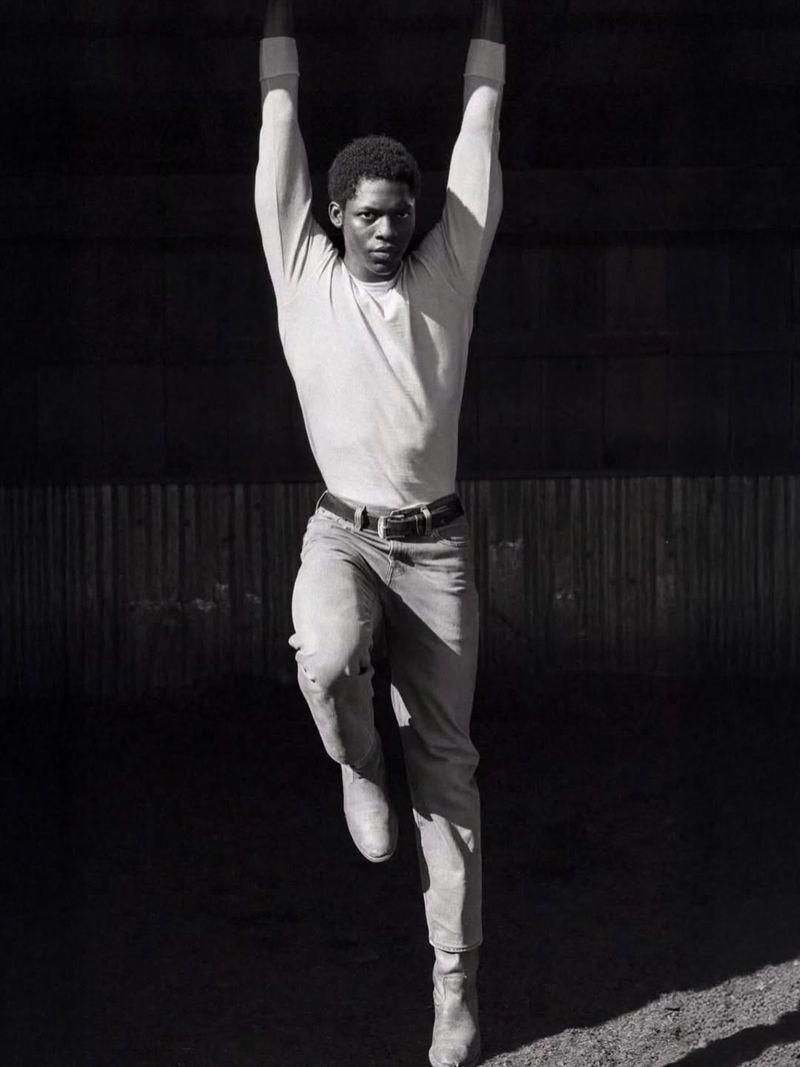 Monochromatic portrait of athletic adult male hanging from bar in boots and jeans against a neutral, shaded indoor backdrop.