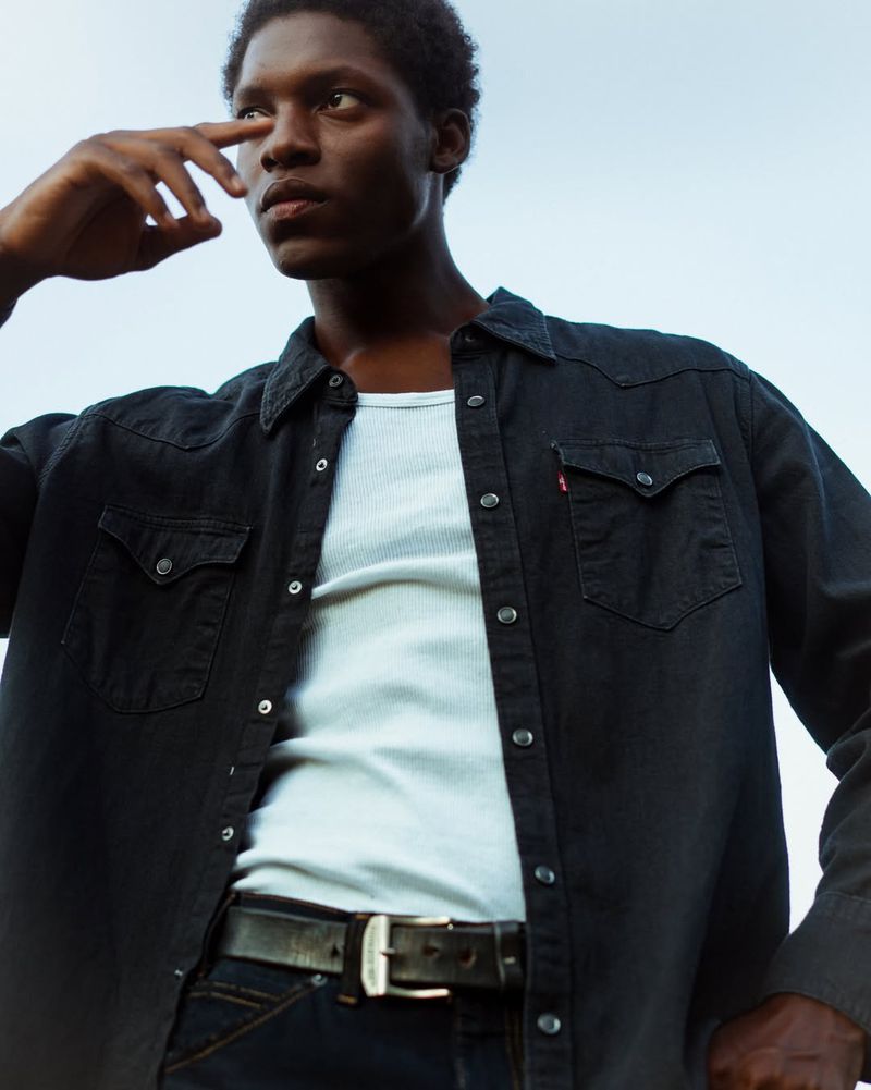 Low-angle shot of a black man wearing an open dark denim shirt over a white ribbed tank top against a clear sky.