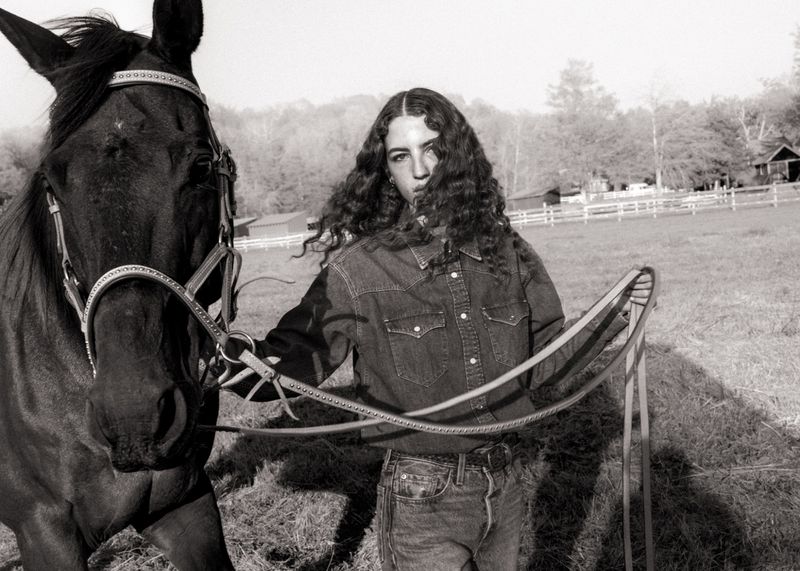 Monochrome portrait of a woman with long dark curly hair wearing a denim shirt standing beside a black horse outdoors.