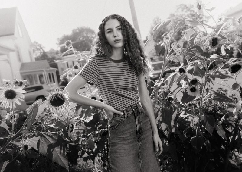 Young woman with long curly hair wearing a striped t-shirt and denim skirt standing among sunflowers outdoors.