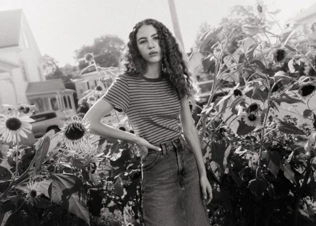 Young woman with long curly hair wearing a striped t-shirt and denim skirt standing among sunflowers outdoors.