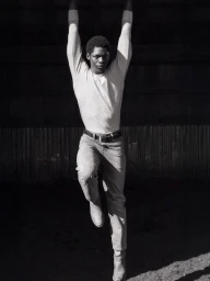 Black man holding overhead with arms raised, wearing long-sleeve top and jeans in monochrome fashion campaign photograph