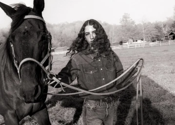 Monochrome portrait of a woman with long dark curly hair wearing a denim shirt standing beside a black horse outdoors.