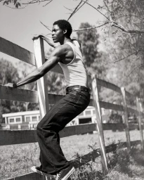 Black man wearing a white tank top and dark jeans climbing a wooden fence outdoors
