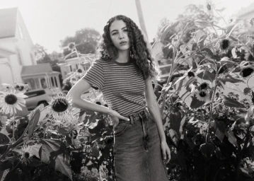 Young woman with long curly hair wearing a striped t-shirt and denim skirt standing among sunflowers outdoors.