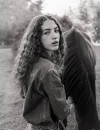 Young woman with long curly hair wearing a denim jacket standing outdoors next to a dark horse in a black and white photograph.