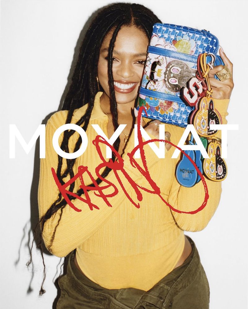 Selah Marley smiling while holding a blue patterned Moynat handbag against a white background