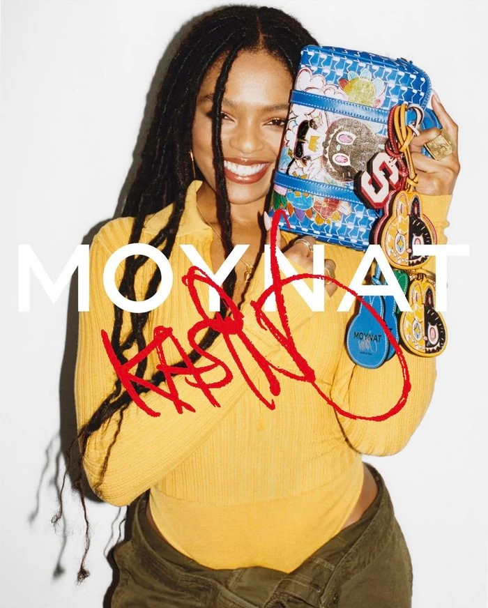 Selah Marley smiling while holding a blue patterned Moynat handbag against a white background
