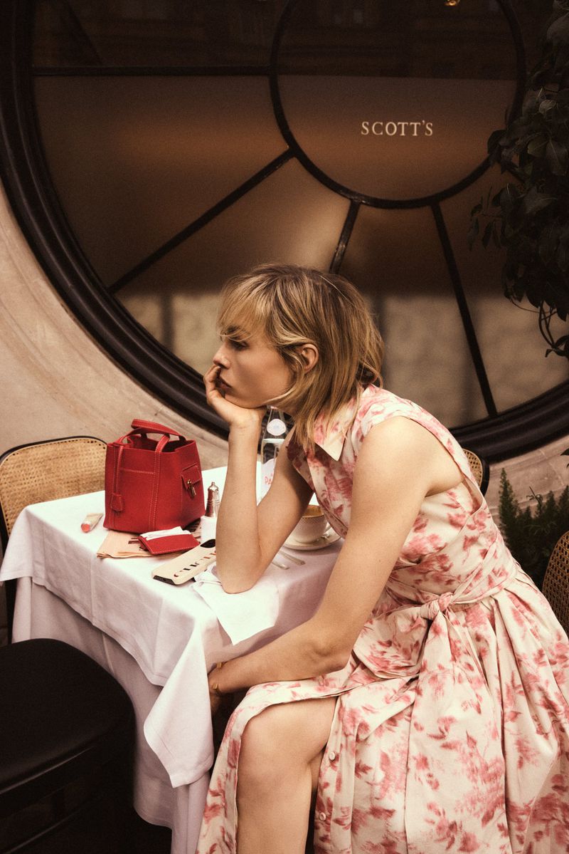 Edie Campbell seated at an outdoor cafe table wearing a pink floral dress beside a red handbag for Carolina Herrera