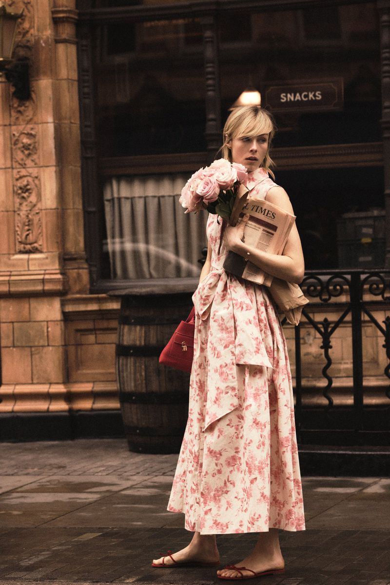 Model Edie Campbell holding pink roses and a newspaper in front of a storefront for Carolina Herrera Matryoshka Pocket.