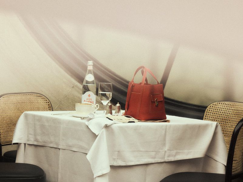 Red leather handbag on a white tablecloth cafe table with a water bottle and glass