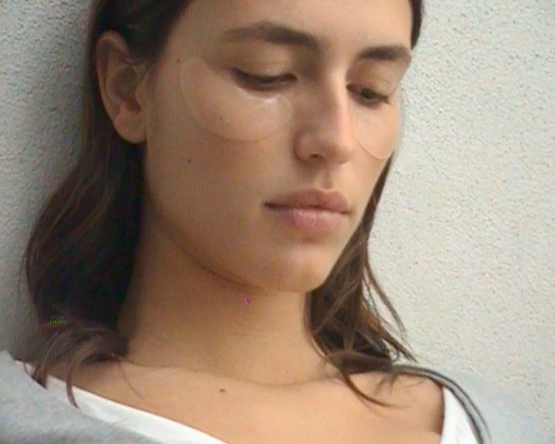 Close-up portrait of a woman with under-eye patches wearing a white top against a textured wall