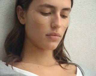 Close-up portrait of a woman with under-eye patches wearing a white top against a textured wall