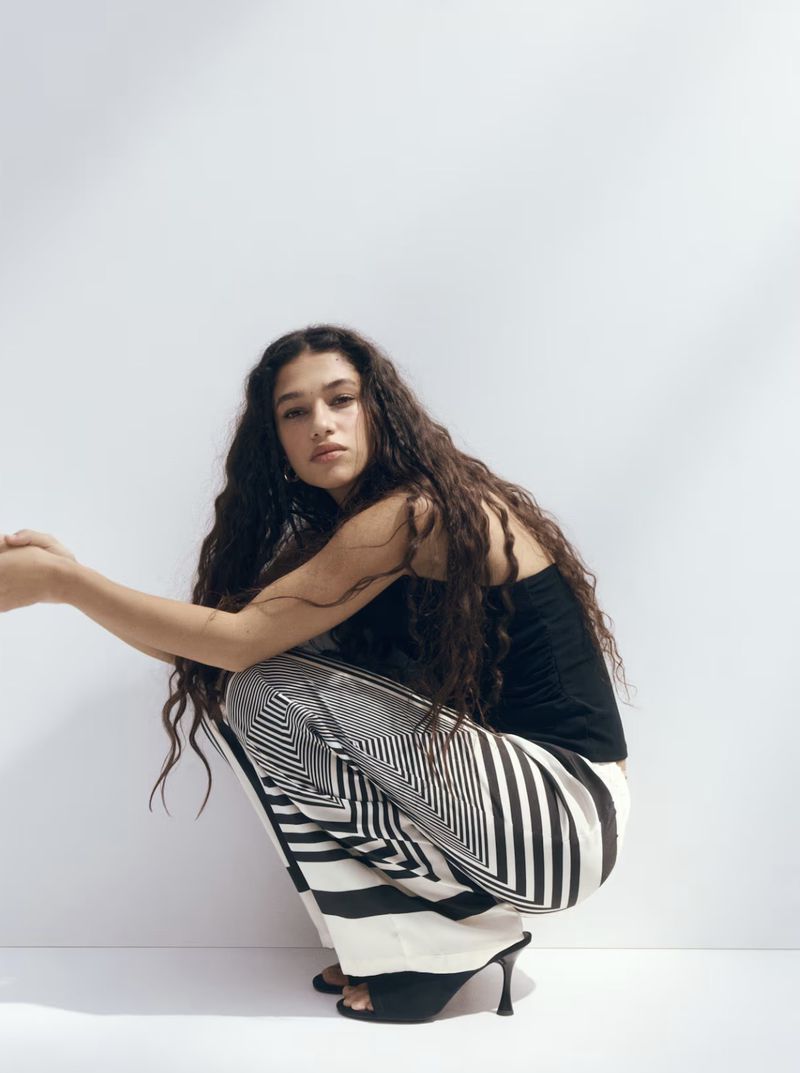 Model with long dark curly hair wearing a black top and patterned trousers crouches against a plain white studio wall.