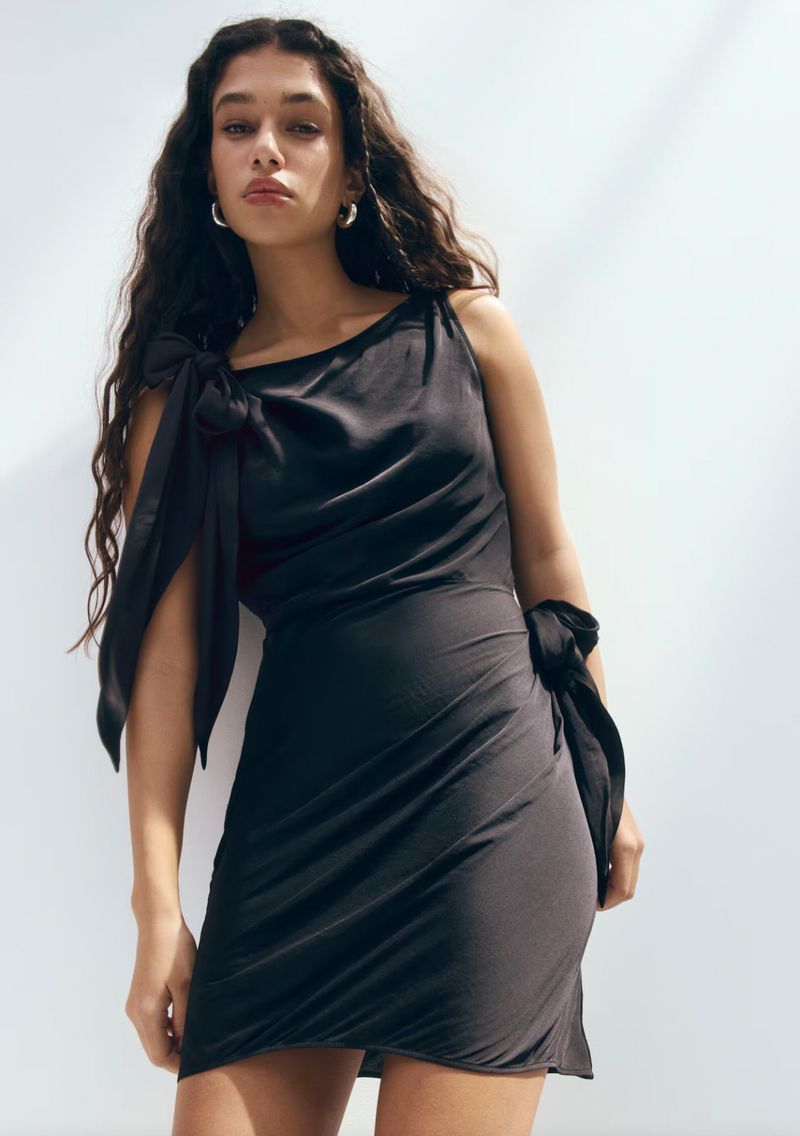 Model wearing a black sleeveless minidress with shoulder and waist ties against a plain white background.