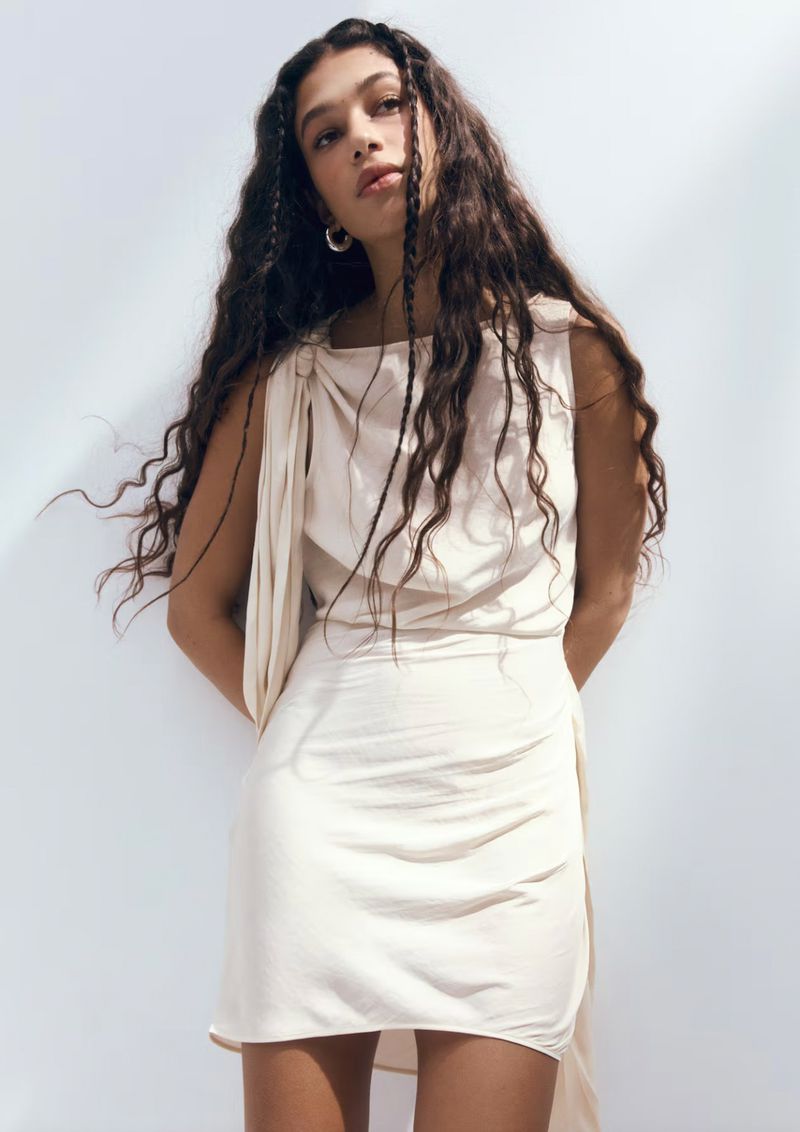 Brunette woman in a sleeveless, light-beige mini dress poses against a plain white backdrop. Her long, wavy hair is styled with small braids.