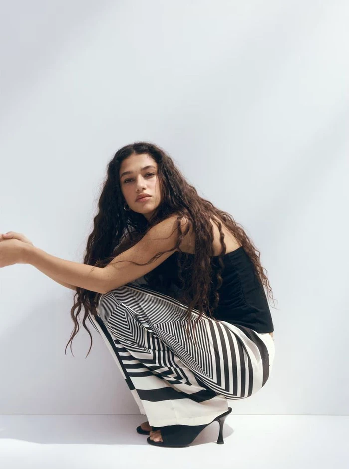 Model with long dark curly hair wearing a black top and patterned trousers crouches against a plain white studio wall.