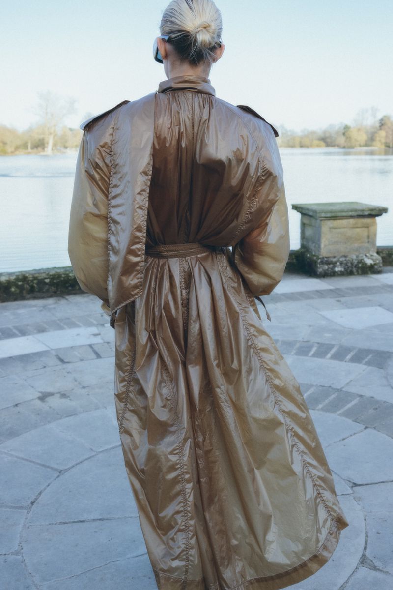 Model seen from behind wearing a glossy brown belted trench coat near a body of water