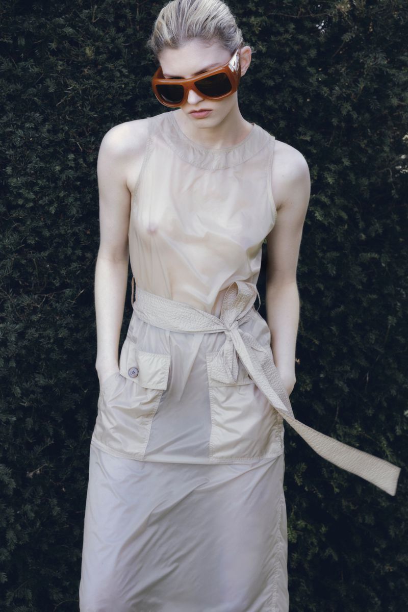 Model wearing oversized sunglasses and a belted neutral dress standing against dark foliage for Net-A-Porter.