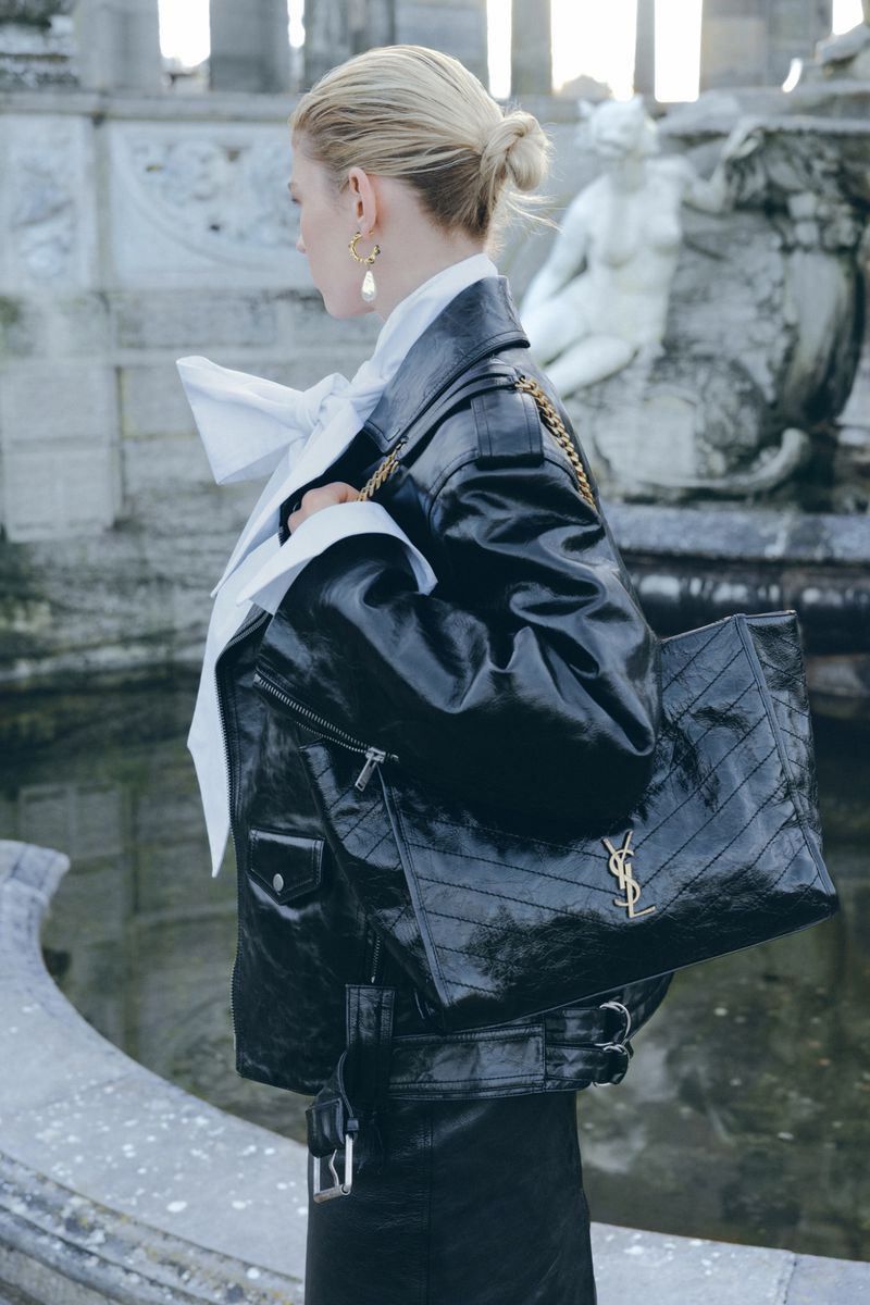 Model in black leather jacket holding a black leather tote bag with gold hardware near stone fountain