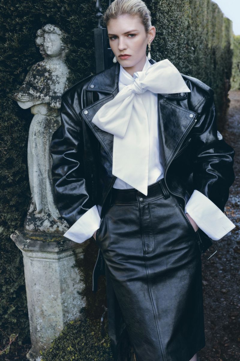 Model wearing a black leather jacket and skirt with a white bow blouse outdoors near a stone statue.