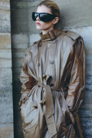 Woman with blonde hair wearing a brown trench coat and black sunglasses posing against a stone wall in a fashion editorial style.
