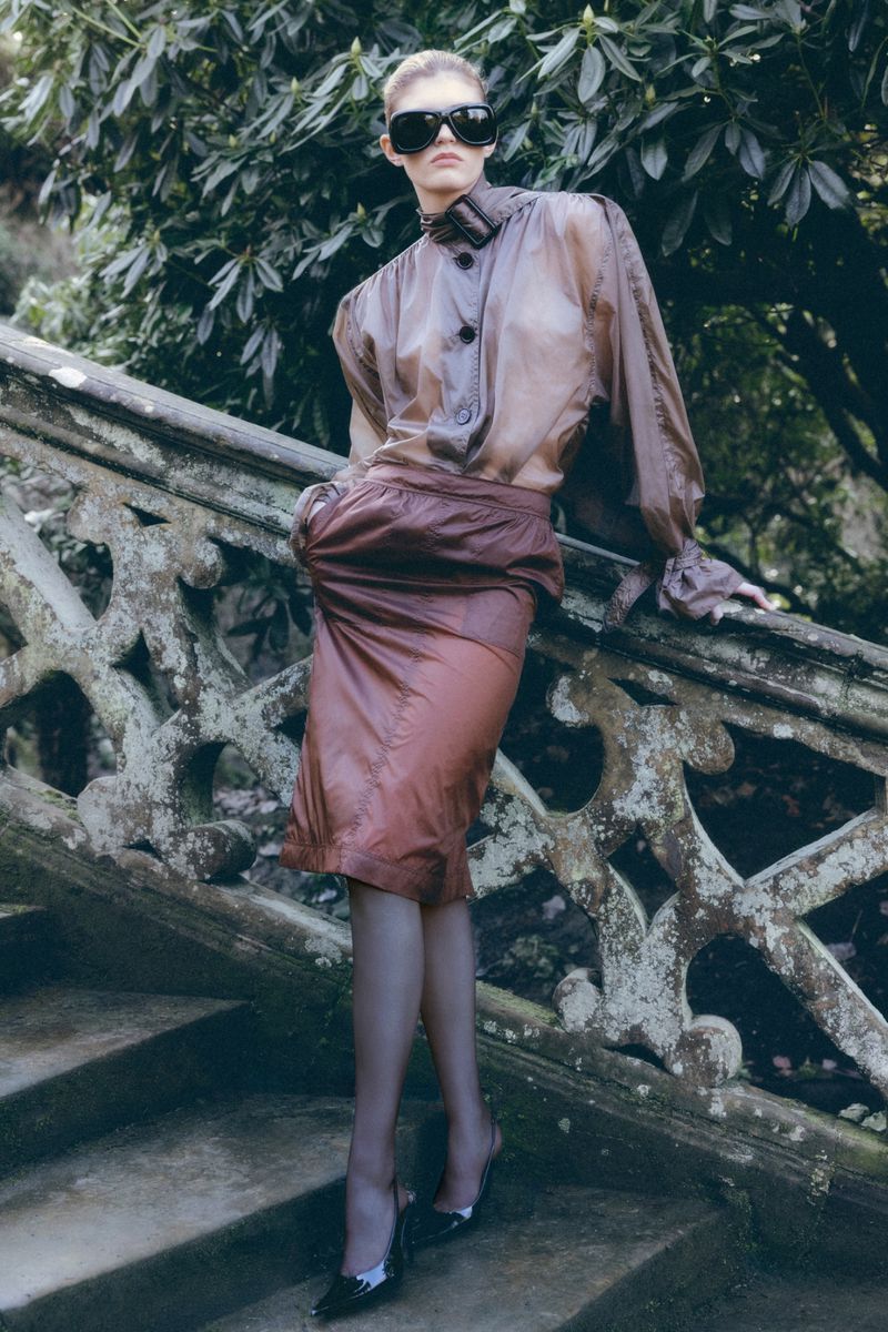 Model in dark oversized sunglasses wearing a sheer brown blouse and skirt leaning against an ornate stone balustrade.