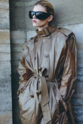 Model in dark sunglasses wearing a long brown belted coat leaning against a stone wall for Saint Laurent