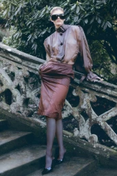 Model in dark oversized sunglasses wearing a sheer brown blouse and skirt leaning against an ornate stone balustrade.