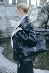Model in black leather jacket holding a black leather tote bag with gold hardware near stone fountain